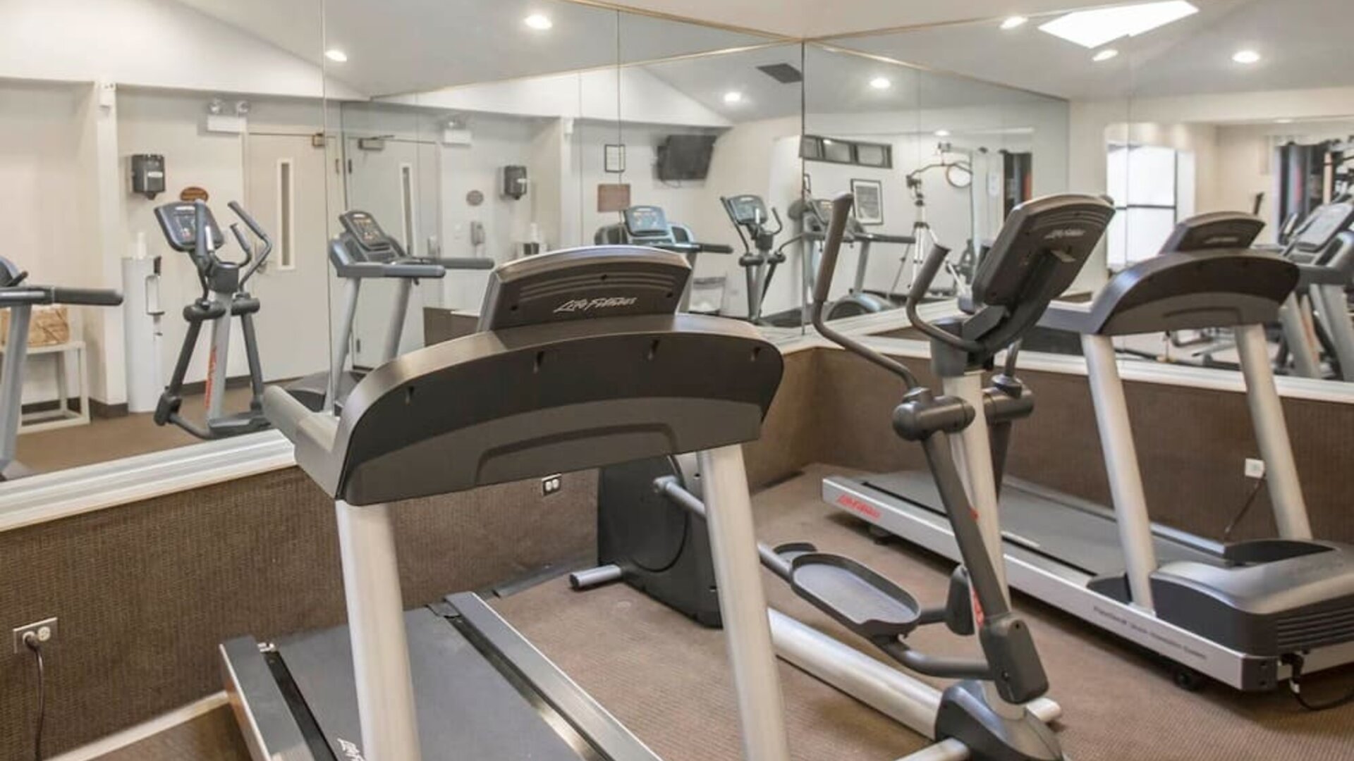 Hotel fitness center featuring multiple treadmills, elliptical exercise machines, wall-to-wall mirrors, bright overhead lighting, and neutral-colored carpeting in a dedicated indoor workout space.
