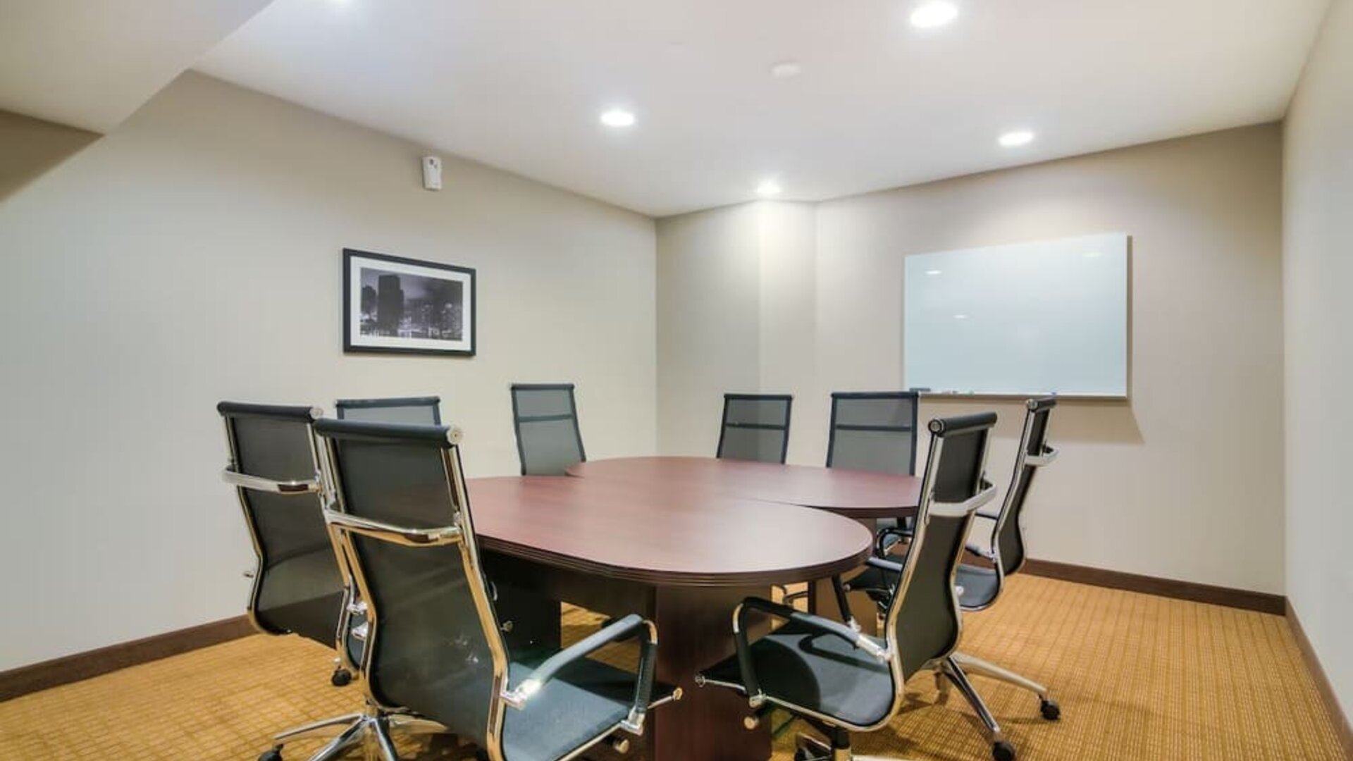Professional hotel meeting room featuring a large oval wooden conference table surrounded by black ergonomic office chairs, a wall-mounted whiteboard, framed artwork, beige carpeting, and recessed ceiling lighting.