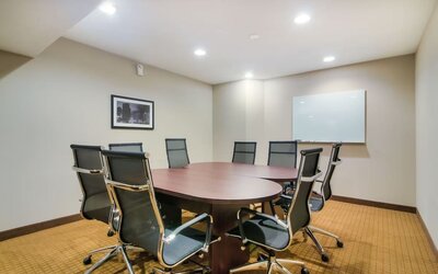 Professional hotel meeting room featuring a large oval wooden conference table surrounded by black ergonomic office chairs, a wall-mounted whiteboard, framed artwork, beige carpeting, and recessed ceiling lighting.