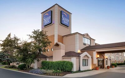 Exterior view of the Sleep Inn hotel at dusk featuring a multi-story beige building, illuminated blue Sleep Inn signage, covered main entrance, landscaped trees and shrubs, decorative lampposts, and a paved driveway.
