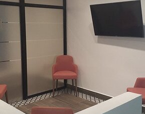 A small lounge area with coral-coloured chairs, a wall-mounted television, tiled flooring and glass partition doors at Residencial & Hotel Palazzo.