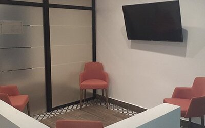 A small lounge area with coral-coloured chairs, a wall-mounted television, tiled flooring and glass partition doors at Residencial & Hotel Palazzo.