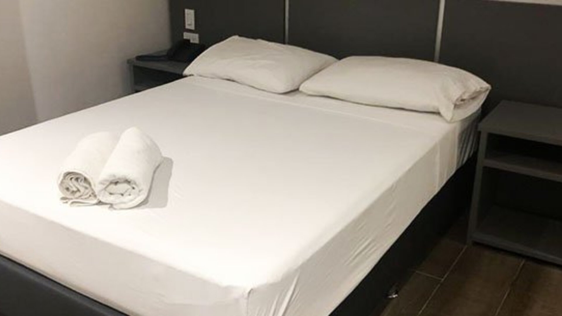 A double bed with white bedding and rolled towels, a dark bedside table with a remote control and a burgundy accent wall at Residencial & Hotel Palazzo.