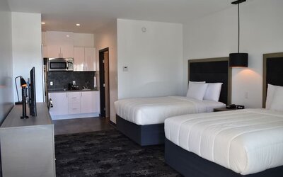 A guest room with two beds dressed in white bedding, black headboards, bedside tables, pendant lighting and a kitchenette in the background at Hotel Bella.