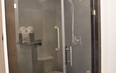 A modern bathroom with a glass-enclosed walk-in shower, dark grey wall tiles, chrome fixtures and a built-in shelf holding folded towels at Hotel Bella.