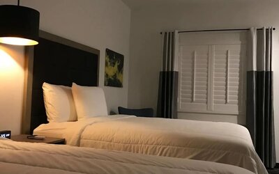A spacious guest room with two beds, white linens, dark bed frames, a television and a compact kitchenette with white cabinetry at Hotel Bella.