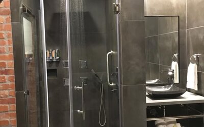 A walk-in shower with glass doors, dark grey tiles, a rainfall showerhead, handheld shower fixture and a wall-mounted shelf at Hotel Bella.