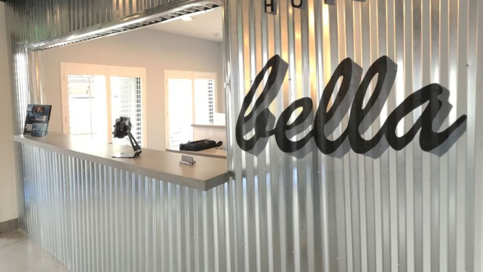 A hotel reception desk set against a corrugated metal wall, featuring a large script-style hotel sign and a bright, minimalist reception area at Hotel Bella.