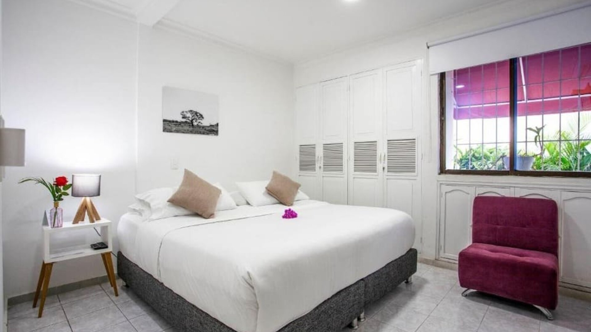 A bright guest room featuring a king-size bed with white linens, beige accent cushions, a magenta upholstered armchair, built-in white wardrobes and a large window overlooking greenery at Mucura Hotel And Spa.