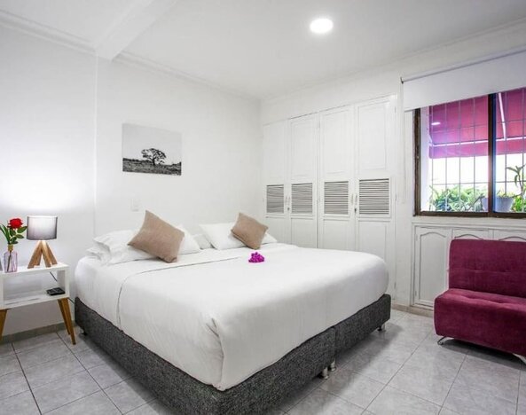 A bright guest room featuring a king-size bed with white linens, beige accent cushions, a magenta upholstered armchair, built-in white wardrobes and a large window overlooking greenery at Mucura Hotel And Spa.