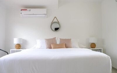 A spacious king bedroom with crisp white bedding, blush and beige accent cushions, matching bedside tables with woven lamps, a round wall mirror and a wall-mounted air conditioner at Mucura Hotel And Spa.