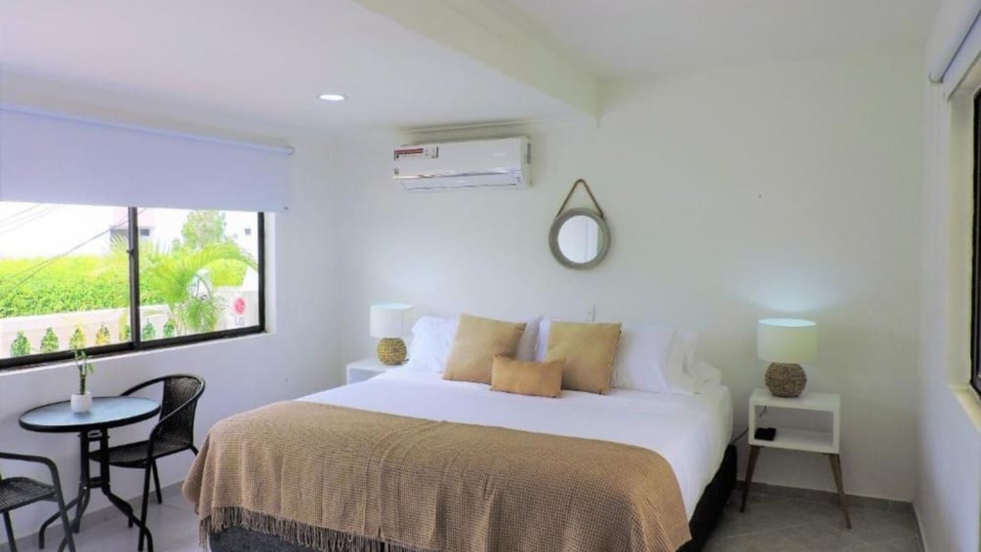 A spacious guest room with a king-size bed dressed in white linens and a beige throw, decorative cushions, matching bedside tables, woven lamps and a small outdoor seating area beside the window at Mucura Hotel And Spa.