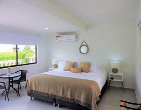 A spacious guest room with a king-size bed dressed in white linens and a beige throw, decorative cushions, matching bedside tables, woven lamps and a small outdoor seating area beside the window at Mucura Hotel And Spa.