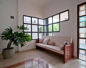 A welcoming lounge area with a wooden-framed sofa upholstered in light fabric, neutral cushions, a large potted plant, tiled flooring and decorative glass-panelled windows at Mucura Hotel And Spa.
