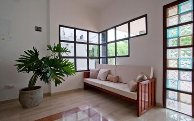 A welcoming lounge area with a wooden-framed sofa upholstered in light fabric, neutral cushions, a large potted plant, tiled flooring and decorative glass-panelled windows at Mucura Hotel And Spa.