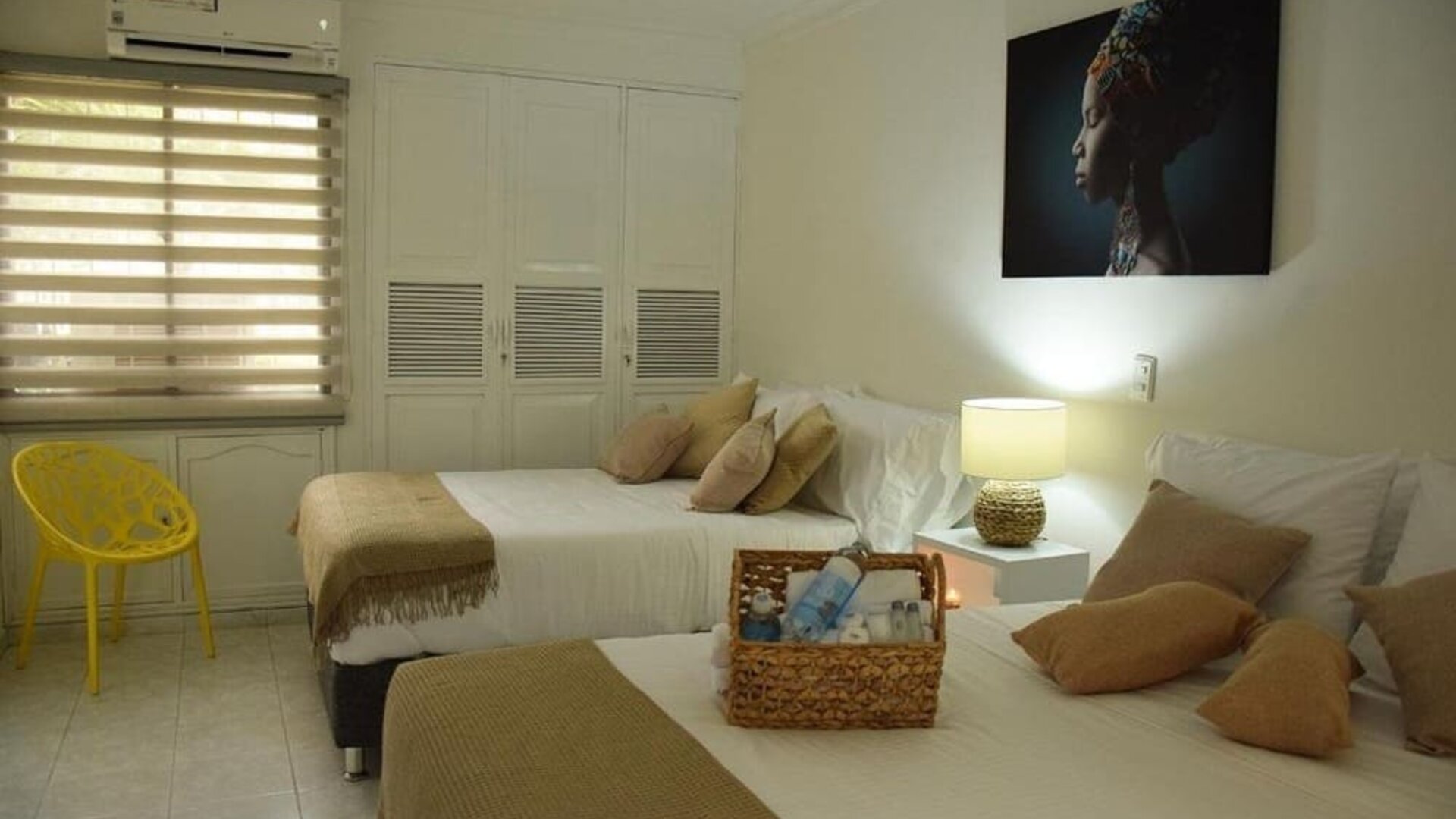A twin-bedroom setup with two beds dressed in white linens and beige throws, decorative cushions, a yellow accent chair, built-in wardrobes and a wall-mounted air conditioner at Mucura Hotel And Spa.