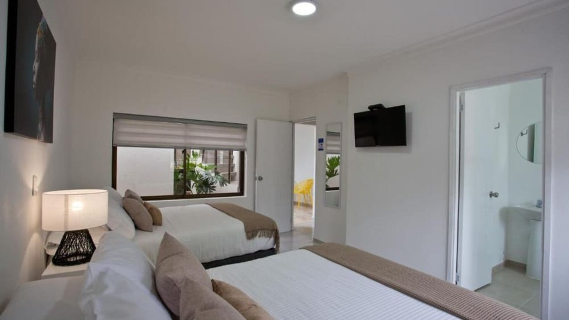A twin-bed guest room with white bedding, beige accent cushions and throws, a bedside table with a decorative lamp, a wall-mounted television and an en-suite bathroom visible through the open doorway at Mucura Hotel And Spa.
