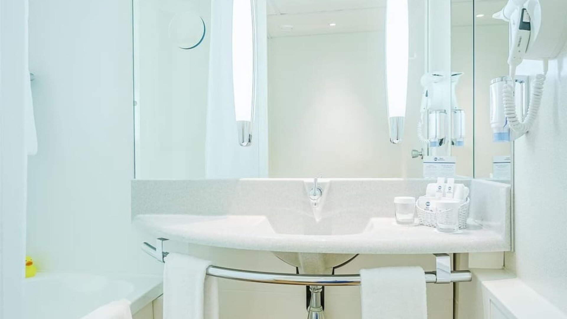 A spacious bathroom with a white bathtub, a curved vanity basin, an expansive mirror, contemporary lighting, a wall-mounted hairdryer and white towels, complemented by warm wood-effect flooring at Best Western Macrander Hotel Frankfurt/Kaiserlei.