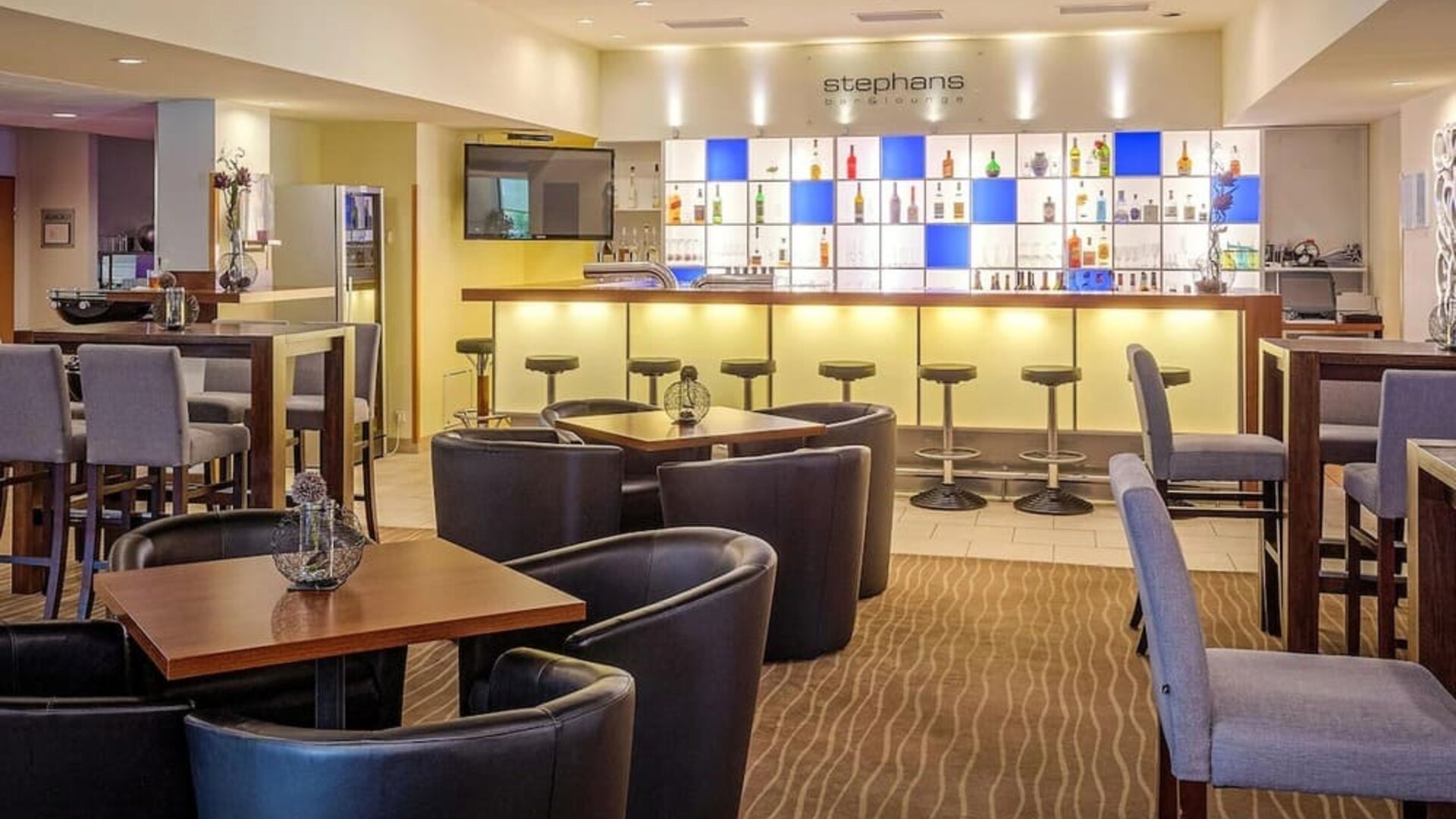 The bar and lounge area at Best Western Macrander Hotel Frankfurt/Kaiserlei, featuring an illuminated backlit bar with a selection of drinks, bar stools, comfortable armchairs, wooden tables and patterned carpeting.