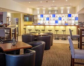 The bar and lounge area at Best Western Macrander Hotel Frankfurt/Kaiserlei, featuring an illuminated backlit bar with a selection of drinks, bar stools, comfortable armchairs, wooden tables and patterned carpeting.