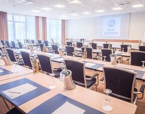 A conference room arranged for meetings and presentations, with rows of tables and chairs, writing materials, water bottles, presentation equipment and a projection screen displaying the Best Western logo.