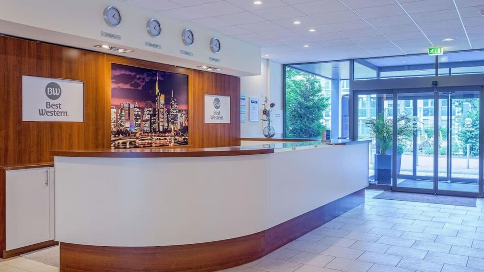 The reception area at Best Western Macrander Hotel Frankfurt/Kaiserlei, featuring a long curved reception desk, wood-panelled walls, hotel branding, wall clocks and glass entrance doors.
