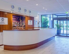 The reception area at Best Western Macrander Hotel Frankfurt/Kaiserlei, featuring a long curved reception desk, wood-panelled walls, hotel branding, wall clocks and glass entrance doors.