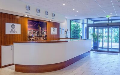 The reception area at Best Western Macrander Hotel Frankfurt/Kaiserlei, featuring a long curved reception desk, wood-panelled walls, hotel branding, wall clocks and glass entrance doors.