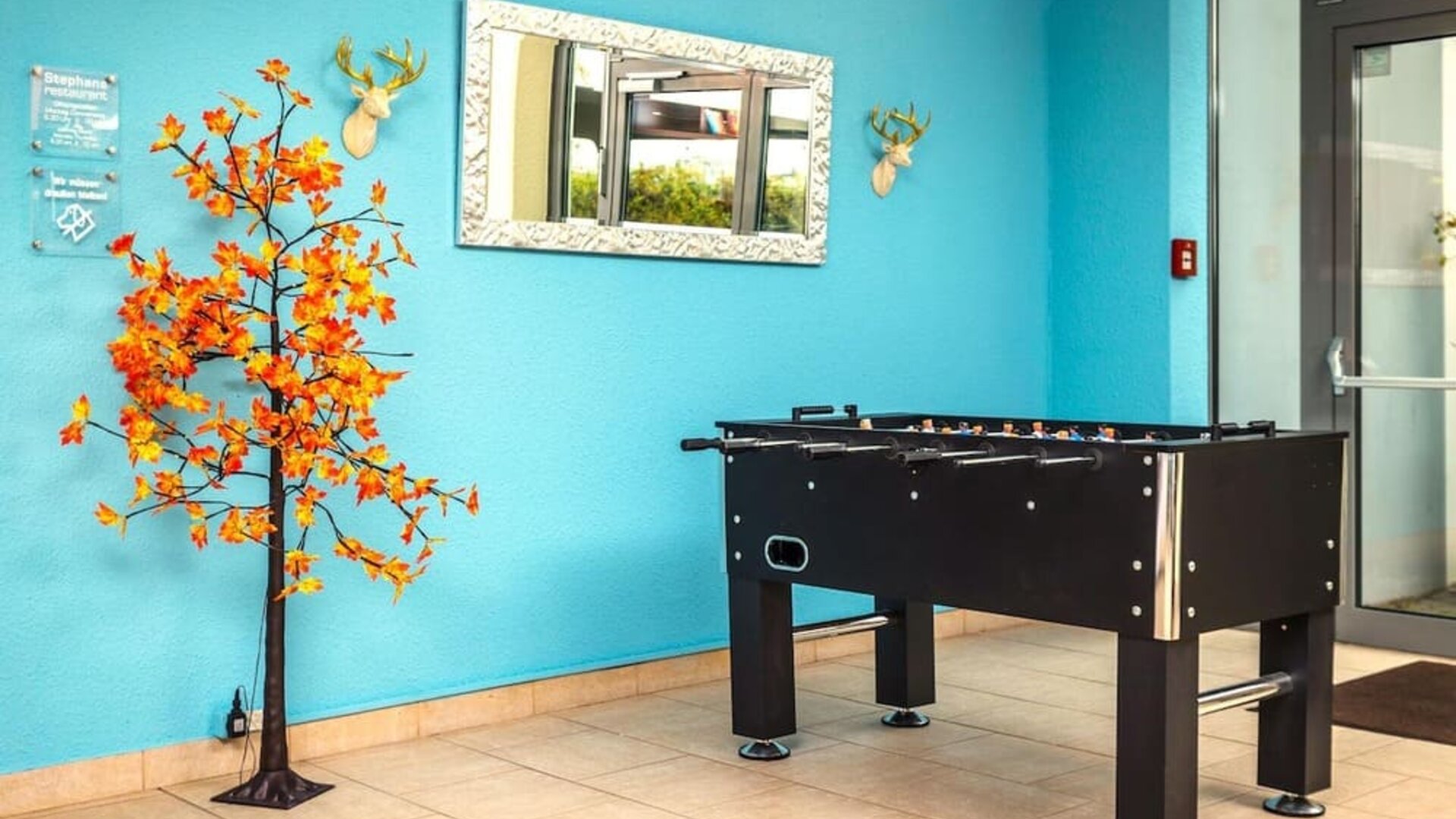 A recreational area featuring a black foosball table against a bright turquoise wall, decorative artwork, a mirror and tiled flooring at Best Western Macrander Hotel Frankfurt/Kaiserlei.