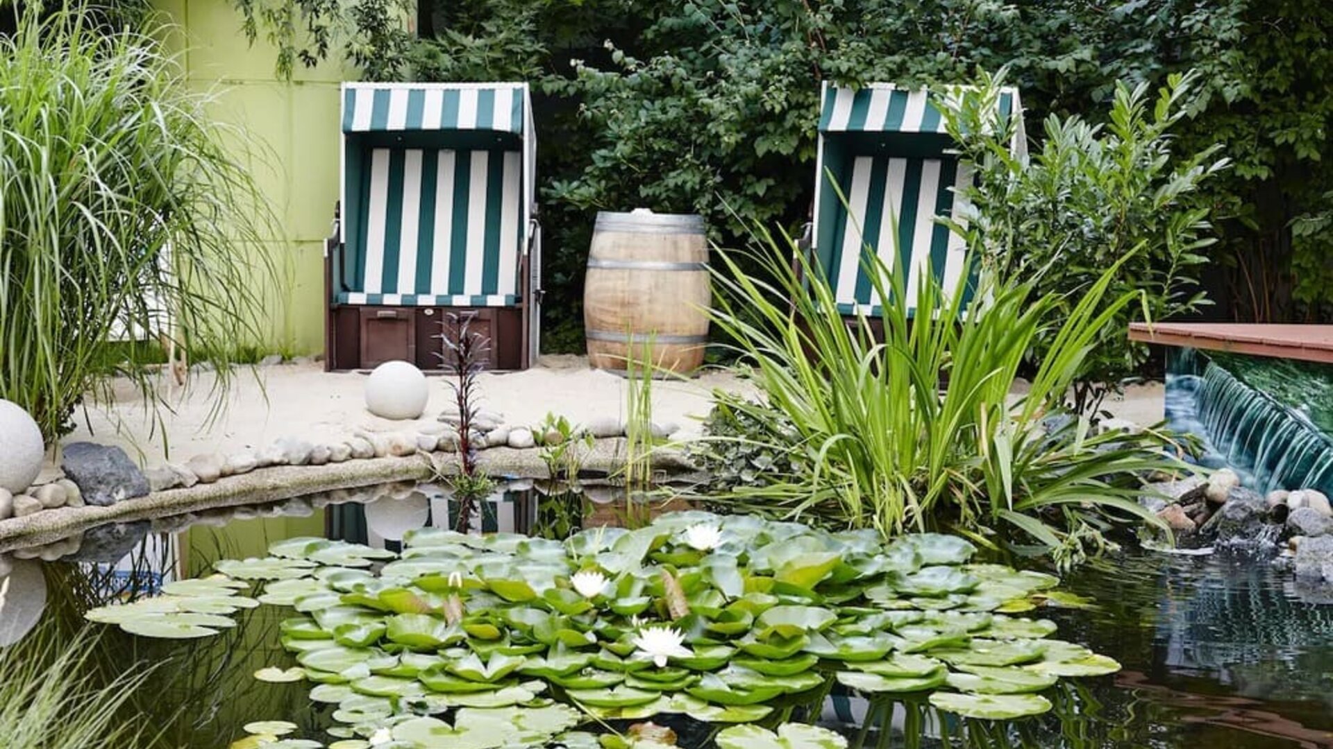 A peaceful landscaped garden with a small pond covered in lily pads and water lilies, surrounded by lush greenery and two striped outdoor deckchairs.