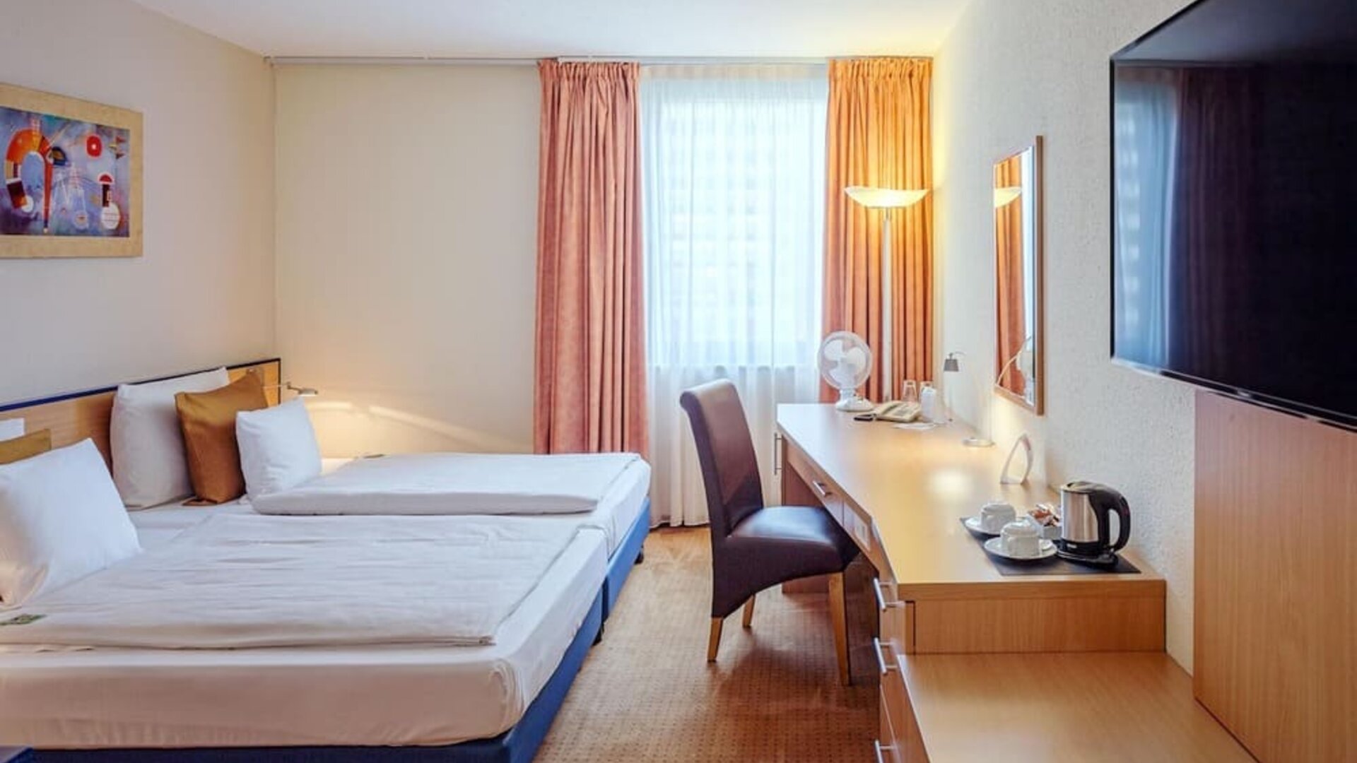Guest room with two single beds dressed in white linens and patterned bedding, warm-toned curtains, a work desk with a chair, a wall-mounted television and a hospitality tray with a kettle and cups at Best Western Macrander Hotel Frankfurt/Kaiserlei.