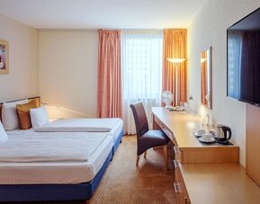 Guest room with two single beds dressed in white linens and patterned bedding, warm-toned curtains, a work desk with a chair, a wall-mounted television and a hospitality tray with a kettle and cups at Best Western Macrander Hotel Frankfurt/Kaiserlei.