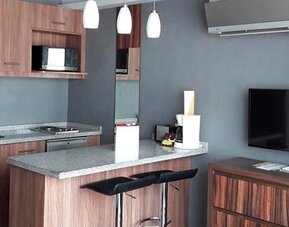 Modern kitchenette and living area with wood cabinetry, microwave, granite breakfast bar, bar stools and wall-mounted television at Hotel Block Suites.