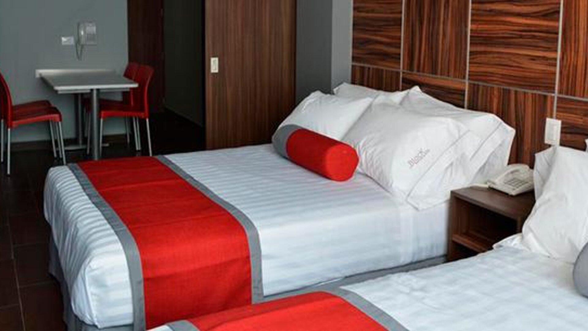 Twin beds with white striped bedding, red runners and cylindrical cushions, dark wood headboard and a small dining table at Hotel Block Suites.