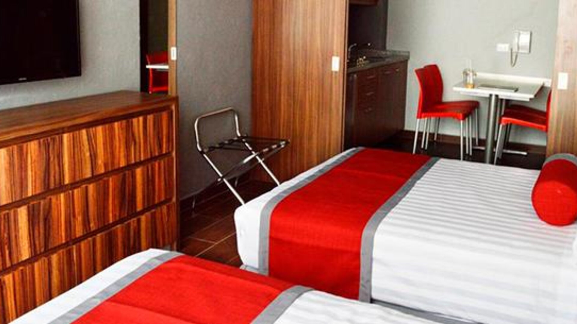 Guest room with two beds featuring white striped bedding, red runners, wood-finish furniture, wall-mounted television and a small dining area at Hotel Block Suites.