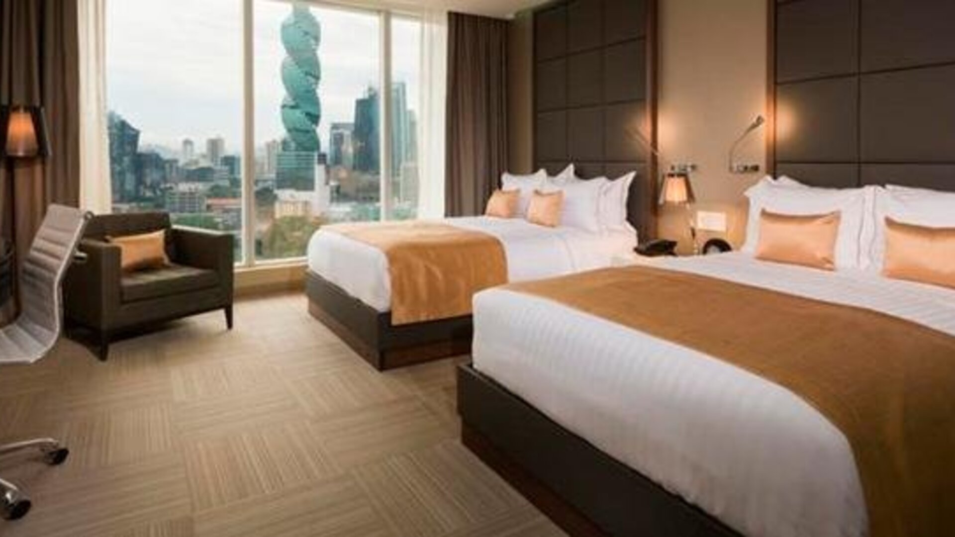 Guest room with two double beds, gold accent cushions, an armchair and city views through large windows at Hotel Las Américas Golden Tower Panamá.