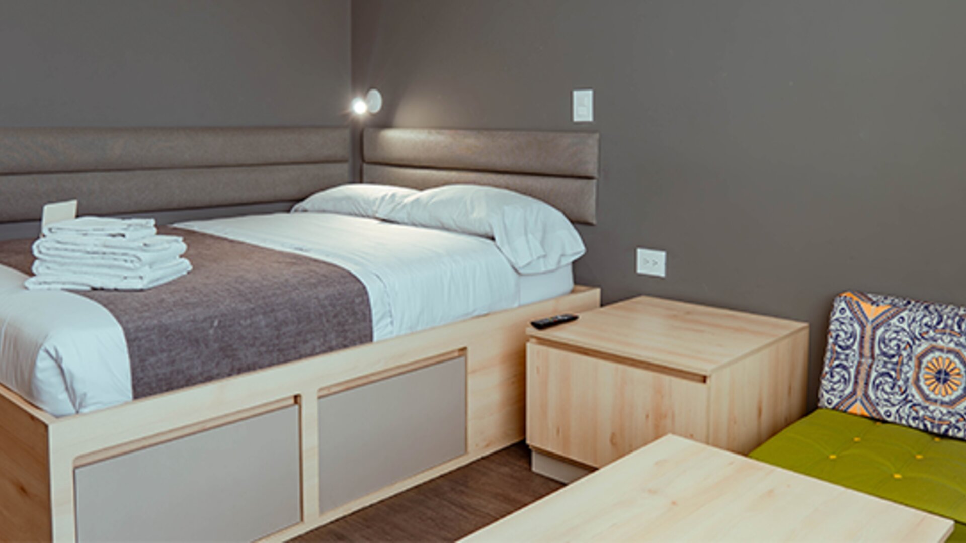 Compact studio with a bed, light wood furniture, upholstered headboard and a green accent chair with a patterned cushion at UMA Suites Chía.