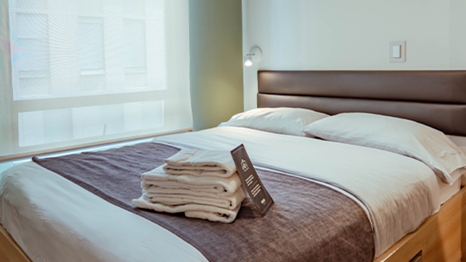 Guest bedroom with a double bed, white bedding, dark grey runner, wooden bed frame and large window with a roller blind at UMA Suites Chía.
