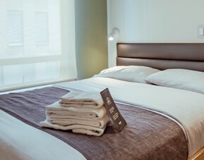 Guest bedroom with a double bed, white bedding, dark grey runner, wooden bed frame and large window with a roller blind at UMA Suites Chía.