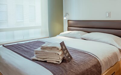 Guest bedroom with a double bed, white bedding, dark grey runner, wooden bed frame and large window with a roller blind at UMA Suites Chía.