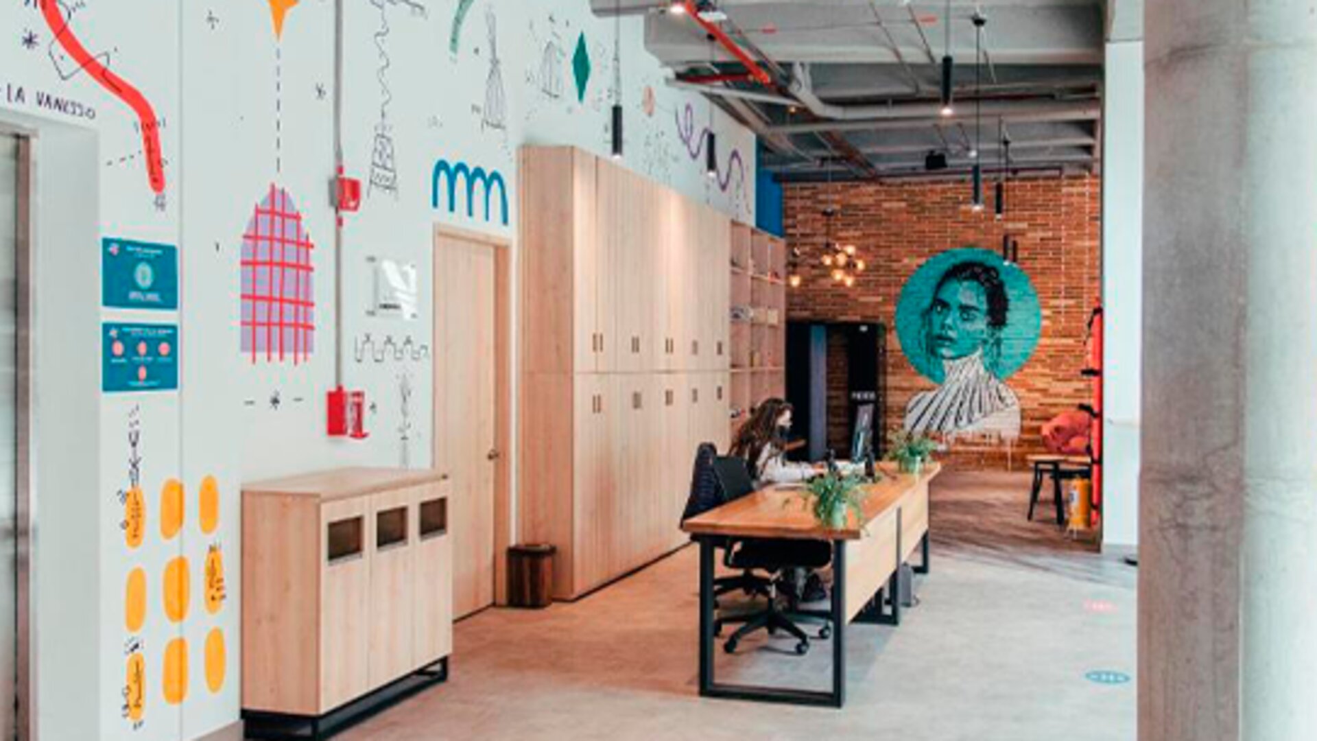 Shared workspace with a long wooden table, chairs, lockers, colourful wall illustrations and exposed concrete columns at UMA Suites Chía.