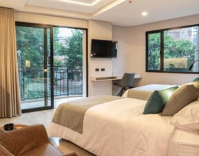 Guest bedroom with two beds, neutral bedding, teal and green accent cushions, a wall-mounted television, desk, and balcony doors at Hotel Living 35 Suites.