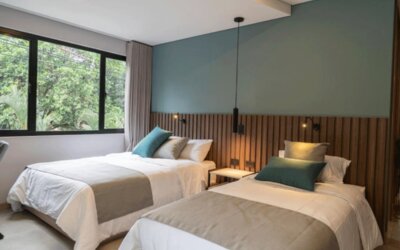 Guest room with two beds dressed in white linens, teal accent cushions, a wooden slat headboard, and large windows at Hotel Living 35 Suites.