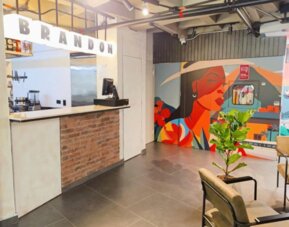 Reception counter with a brick-front desk, illuminated BRANDON lettering, colourful mural artwork, and indoor plants at Hotel Living 35 Suites.