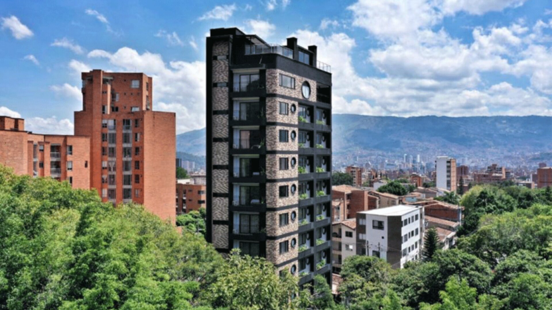 Exterior view of a multi-storey dark-brick hotel building surrounded by green trees, with city buildings and mountains in the background at Hotel Living 35 Suites.