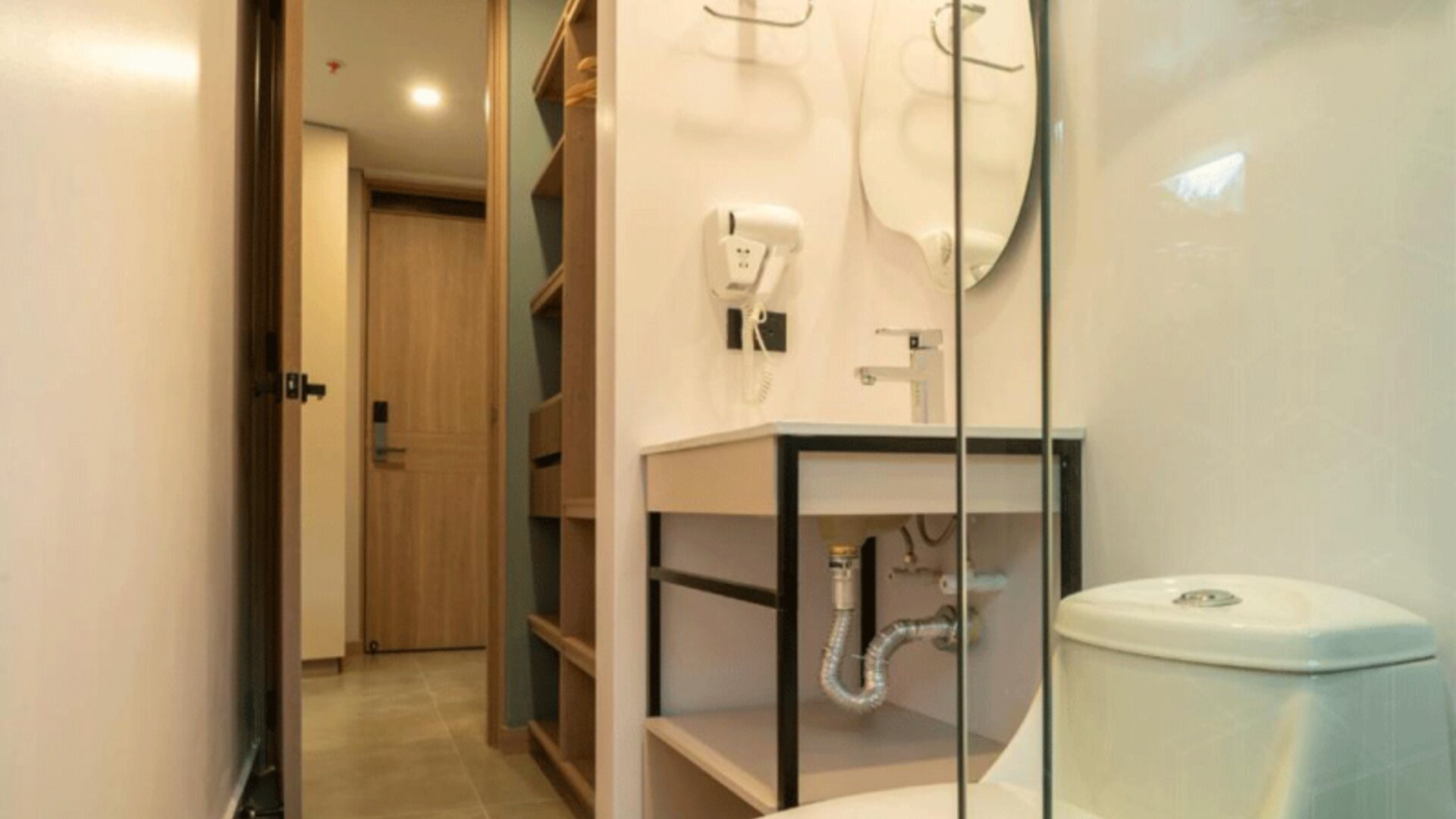 Compact bathroom with a white toilet, open-shelf vanity, wall-mounted hairdryer, glass shower partition, and beige tiled flooring at Hotel Living 35 Suites.