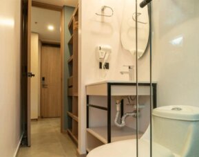 Compact bathroom with a white toilet, open-shelf vanity, wall-mounted hairdryer, glass shower partition, and beige tiled flooring at Hotel Living 35 Suites.