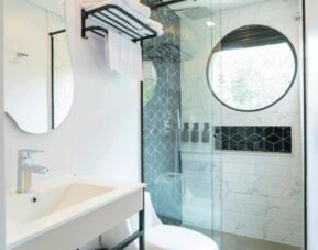 Modern bathroom with a white vanity, curved mirror, glass-enclosed shower, white and grey patterned tiles, and a round window at Hotel Living 35 Suites.