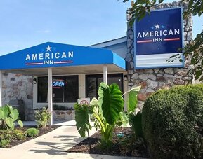Exterior entrance of American Inn Carlisle featuring a blue entrance canopy, hotel signage, stone façade, landscaped plants, and shrubs.