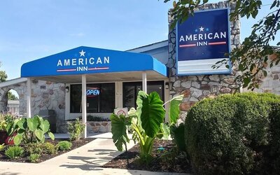 Exterior entrance of American Inn Carlisle featuring a blue entrance canopy, hotel signage, stone façade, landscaped plants, and shrubs.
